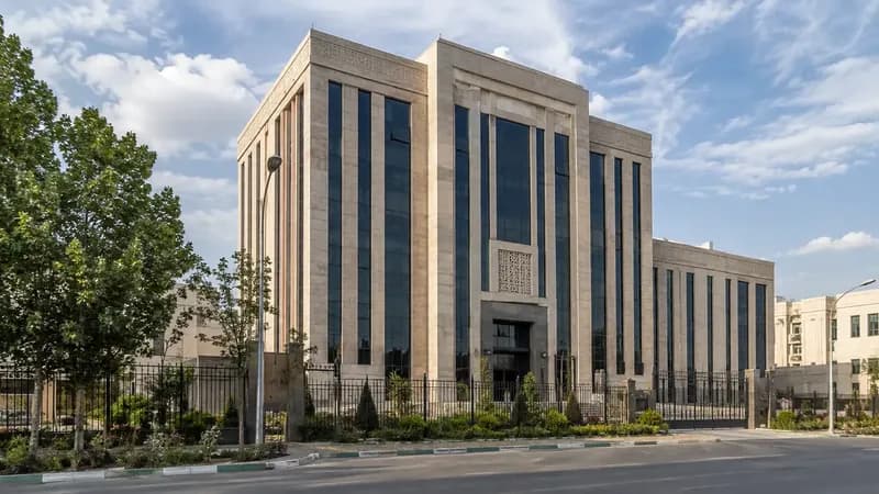 ساختمان اداری بانک با نمای سنگی و ستونهای شیشهای آبی و درختان مقابل آن